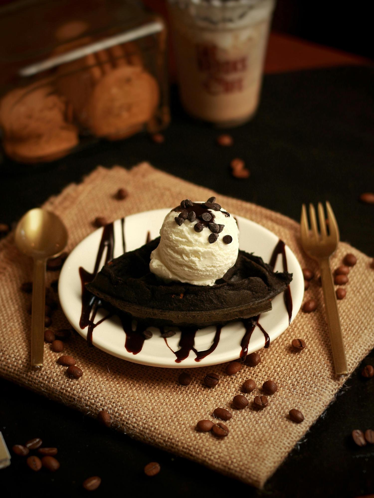 A dark waffle with a scoop of ice cream and chocolate sauce is centered on a white plate, set on a burlap surface scattered with coffee beans, with a gold spoon and fork to either side. Other desserts are softly visible in the background.