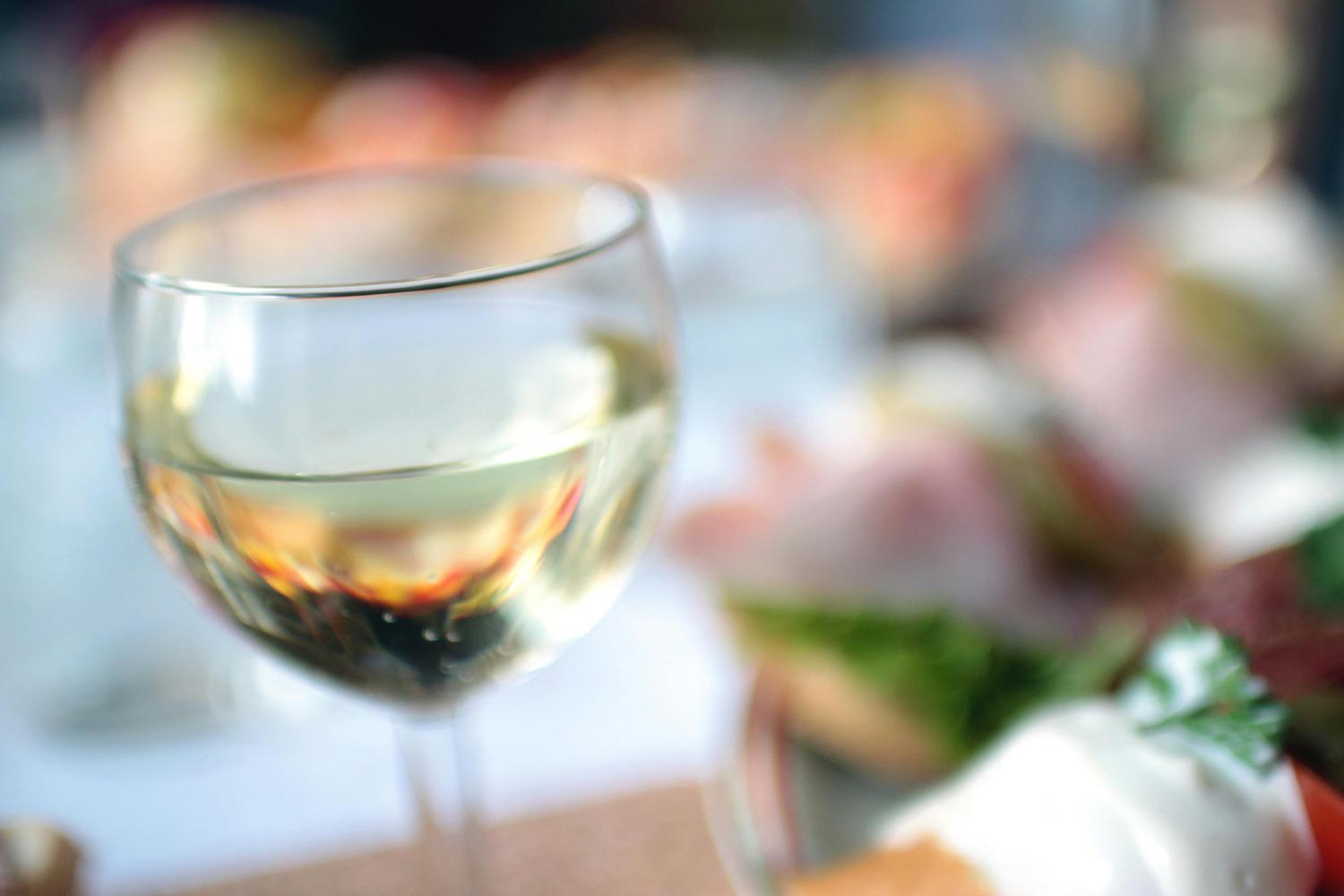 Close-up of a glass of white wine with appetizers blurry in the background. The appetizers include tomatoes, ham, and parsley.