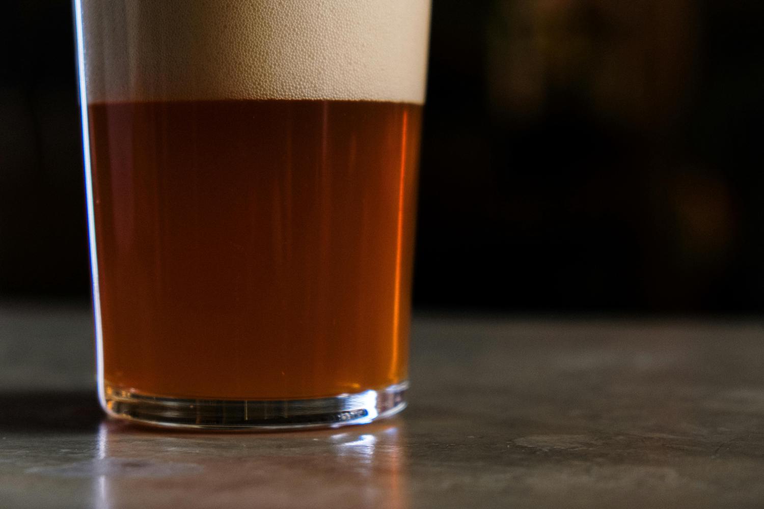 A close-up of a pint glass filled with amber beer, topped with a thick, foamy head, resting on a textured surface against a dark background. The lighting highlights the beer's color and the texture of the foam.