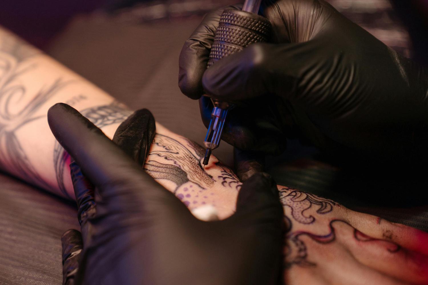 Close-up shows a tattoo artist wearing black gloves, tattooing a design on a client's forearm. The client already has other tattoos.