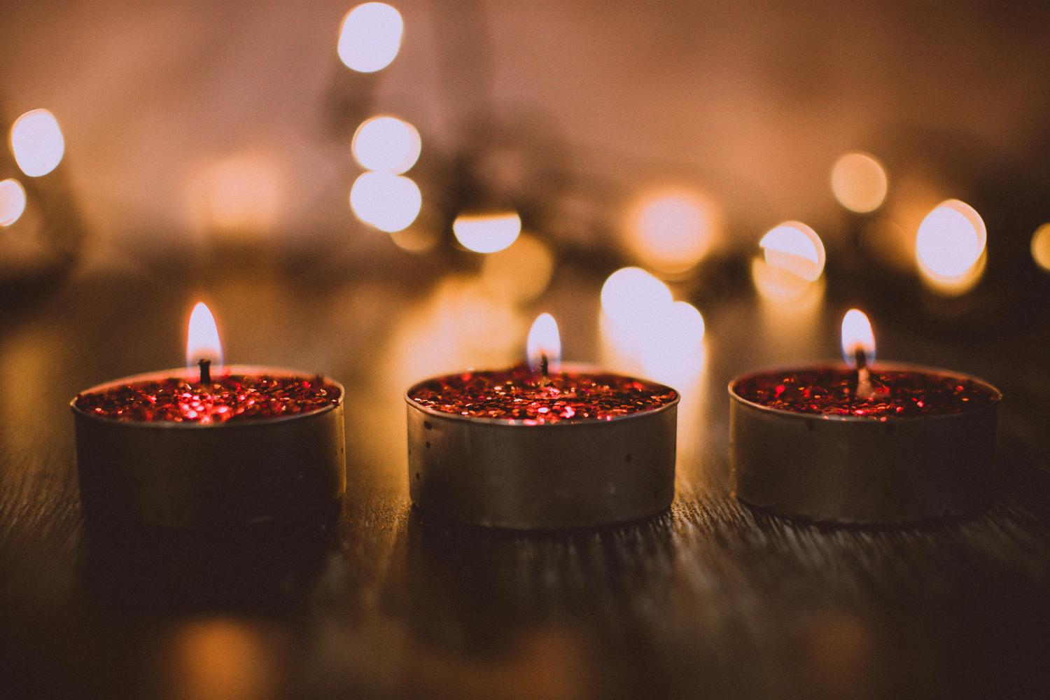 Three lit candles with red glitter sit on a wooden surface with soft, blurred lights in the background. The candles create a warm and festive atmosphere.