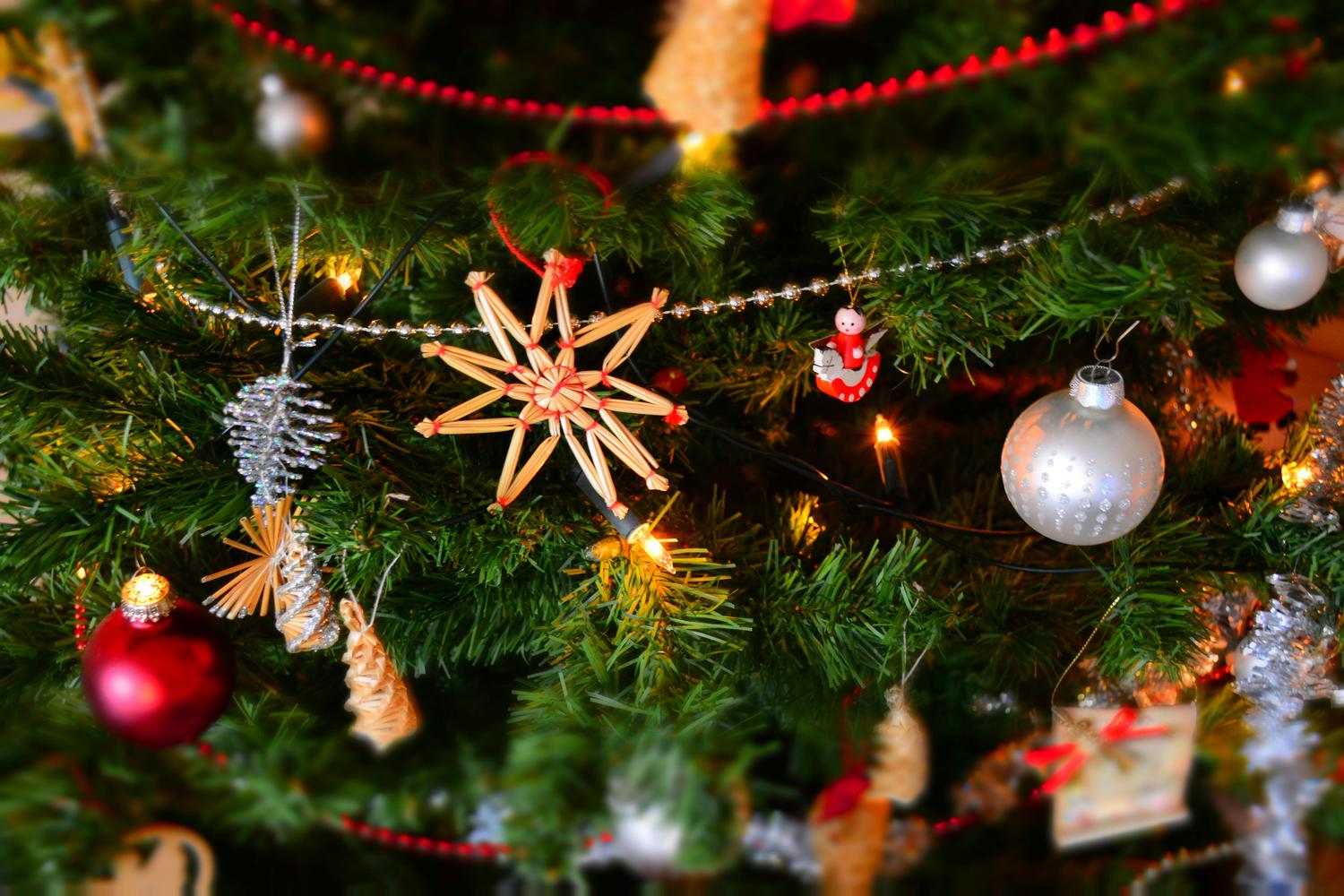 Close-up of a decorated Christmas tree featuring ornaments like a straw star, silver pinecone, and red bauble. A string of lights adds a warm glow to the festive scene.