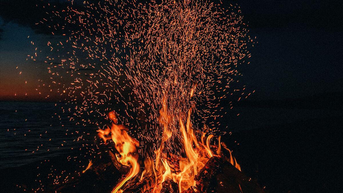 A bonfire burns against a dark, twilight sky with the sea visible on the left; sparks fly upwards in a shower of light.