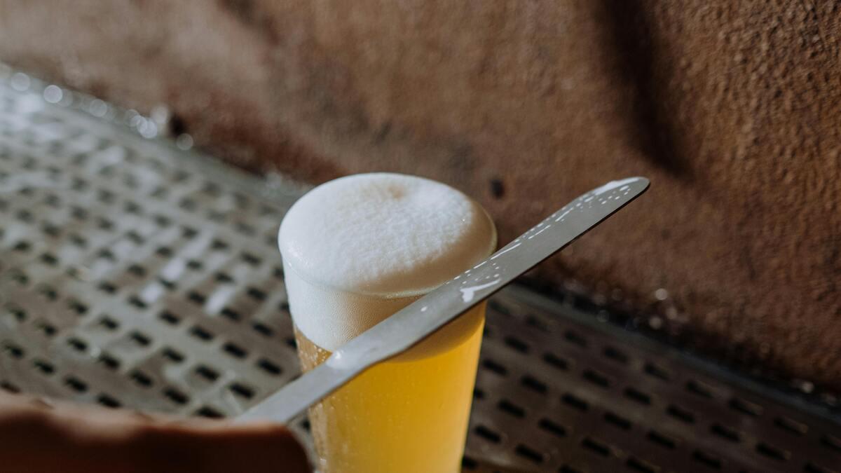 A glass of beer with a thick, foamy head rests on a metal surface, and a knife is being used to trim the foam. The background features a textured, dark brown surface.