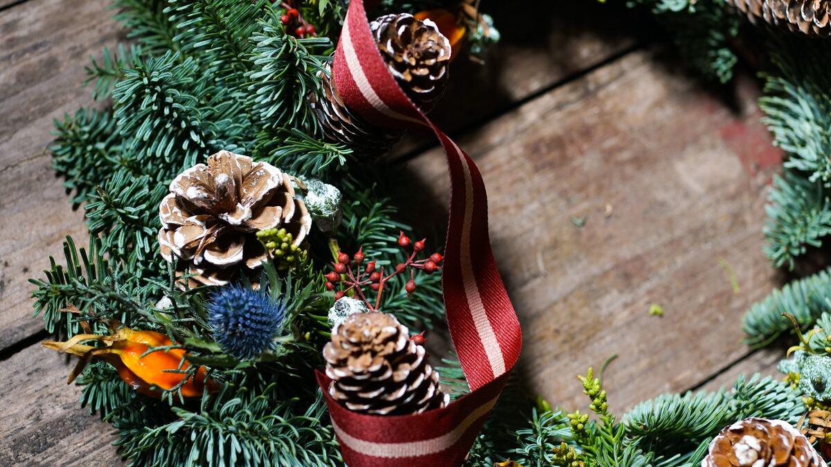 A decorated Christmas wreath rests on a weathered wooden surface, featuring evergreen branches, pine cones, berries, and a striped ribbon. The arrangement creates a festive holiday display.