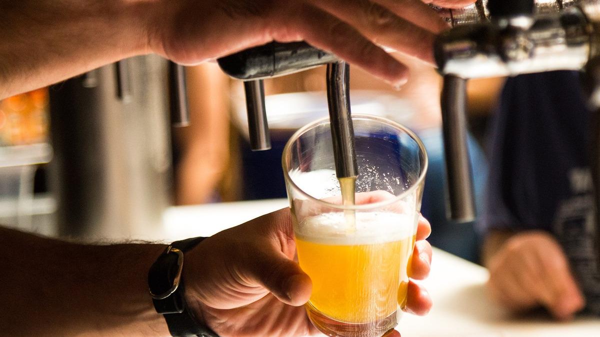 Close-up of a hand pulling a beer tap and filling a glass with golden beer and white foam. The hand wearing a watch steadies the glass beneath the tap.