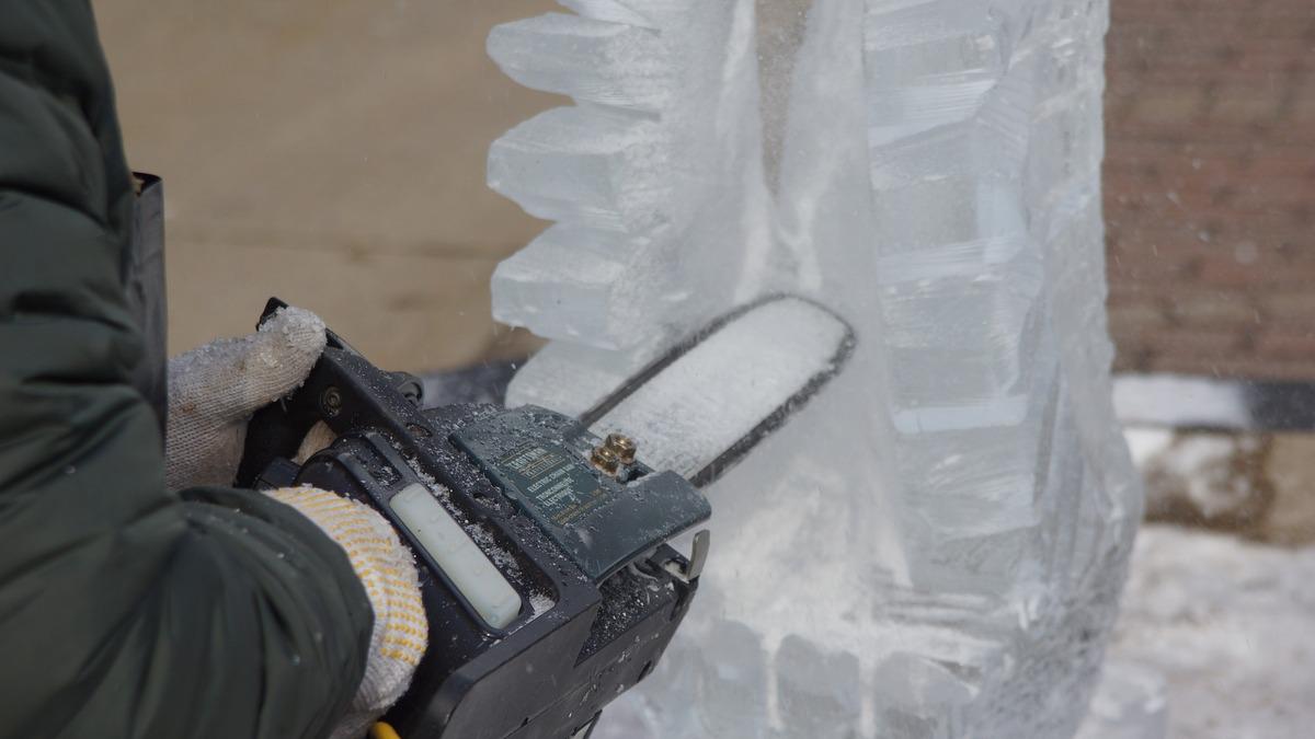 Close-up shows an ice sculptor using a chainsaw to shape a large block of ice. The artist wears gloves and a jacket.