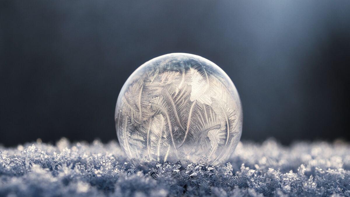 A frozen soap bubble rests on a bed of icy crystals, the bubble's surface covered in intricate frost patterns resembling ferns and feathers against a dark background. The light reflects off the ice crystals, creating a shimmering effect.