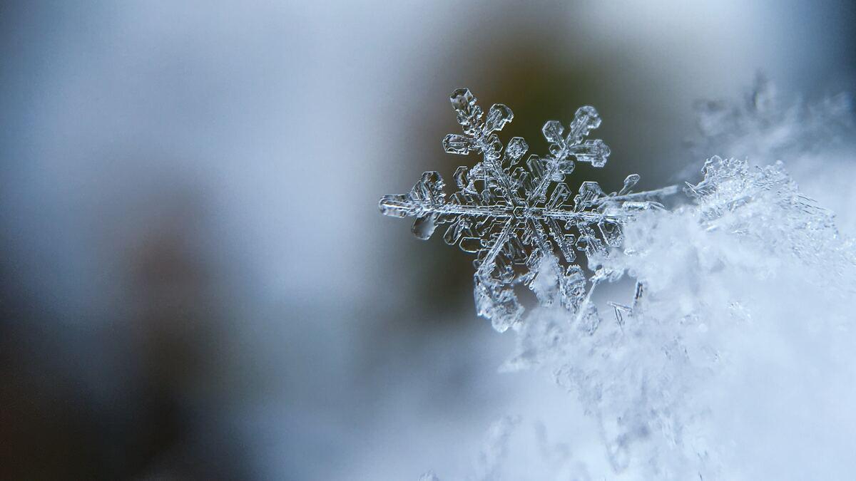 A crystalline snowflake is captured in close-up against a soft, blurred background, resting on a bed of other snowflakes. The snowflake exhibits intricate, symmetrical details.