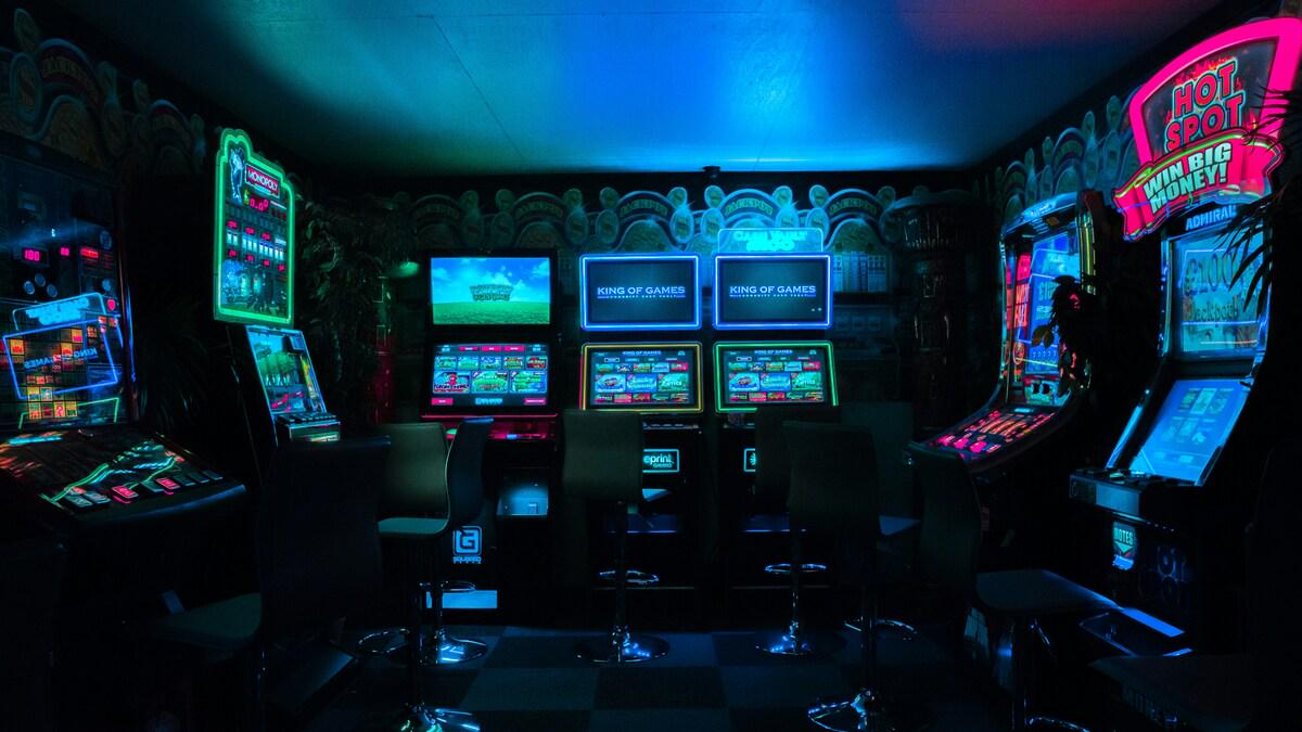 Dimly lit casino interior featuring rows of glowing slot machines in various colors and designs, with empty stools in front of them. The room has a retro aesthetic with decorative wall details.