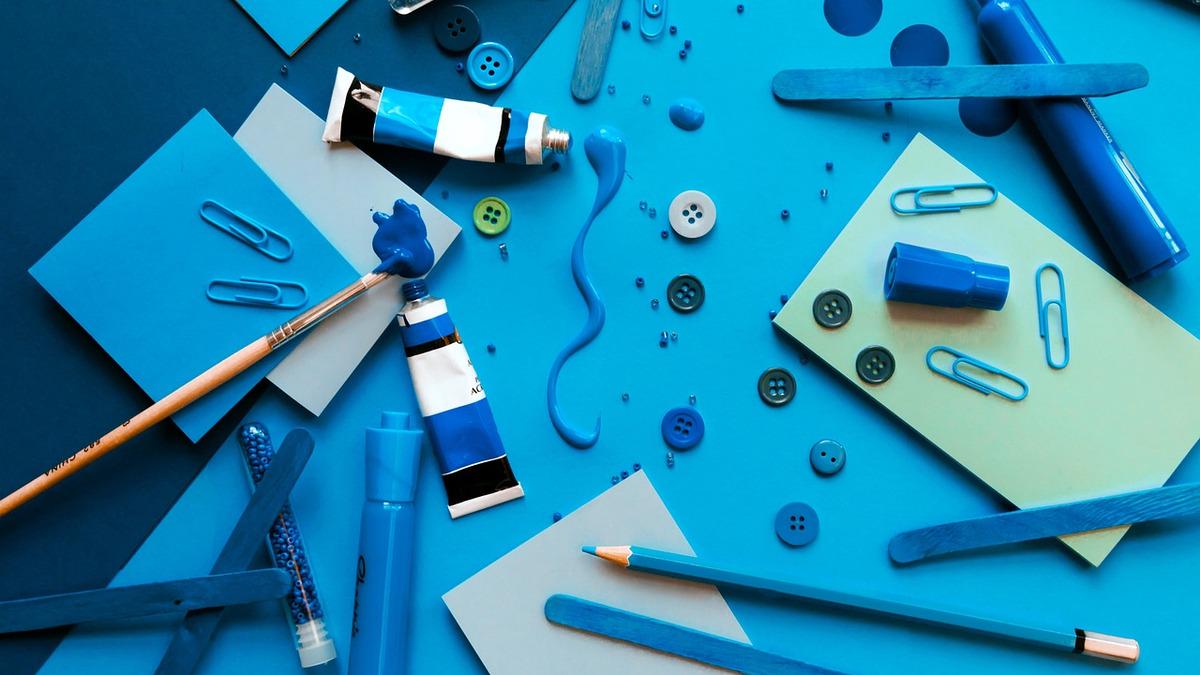 A still-life arrangement of art and craft supplies in various shades of blue are spread across a blue background, including paint tubes, a paintbrush, buttons, paperclips, and small wooden sticks. A squiggly line of blue paint is also present.