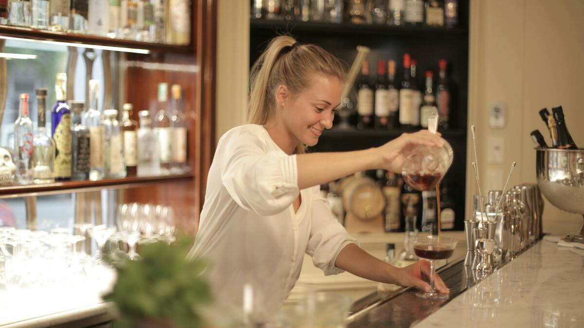 A smiling bartender in a white blouse pours a cocktail from a glass decanter into a stemmed glass, with barware and liquor bottles displayed in the background. The scene is set in a well-lit, upscale bar.
