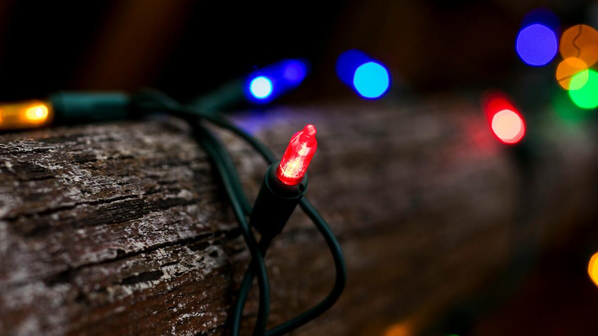 Close-up of a strand of green Christmas lights with red, blue, yellow, and green bulbs, draped over a rough, weathered wooden surface. A red bulb in the foreground is in focus, with others blurred in the background.