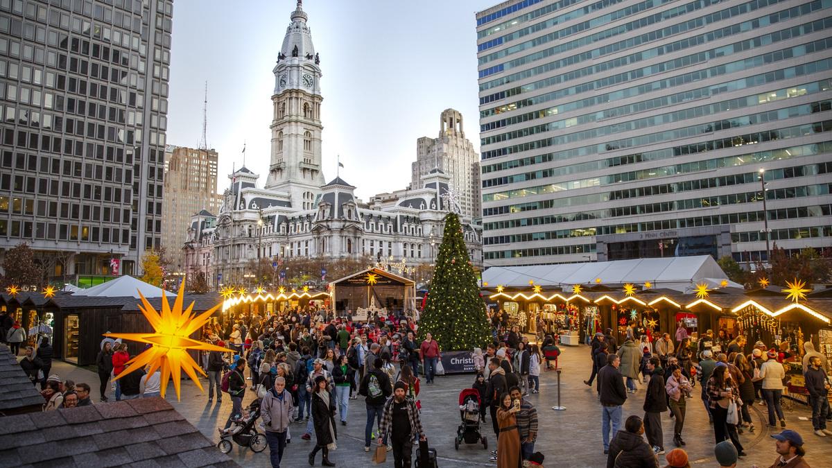 A bustling Christmas Village in Philadelphia's Dilworth Park, featuring wooden stalls, a large Christmas tree, and the iconic City Hall in the background. Crowds of people stroll through the market, enjoying the festive atmosphere.