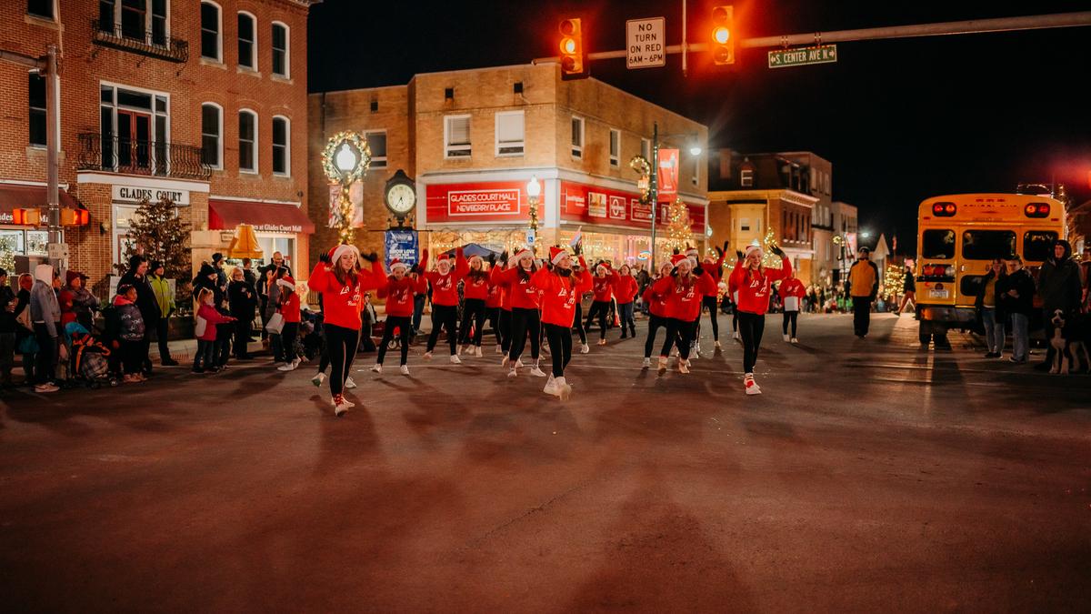 A group of dancers in matching red sweatshirts and Santa hats perform on a street at night, illuminated by streetlights and Christmas decorations, with spectators and a yellow school bus visible in the background. The event is happening in front of brick buildings adorned with holiday wreaths and signs.