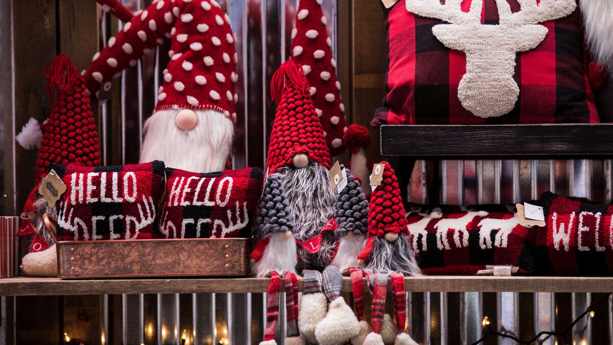A shelf displays a variety of red and black holiday-themed decor, including gnomes with knitted hats, cushions featuring moose designs, and pillows with the words 