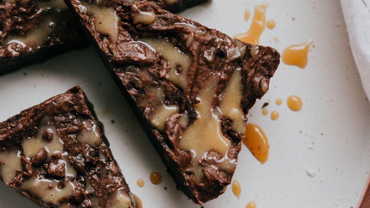 Close-up of caramel-covered brownie pieces arranged on a white surface with scattered drips of caramel sauce. The brownies have a dark chocolate base and are sprinkled with coarse salt.