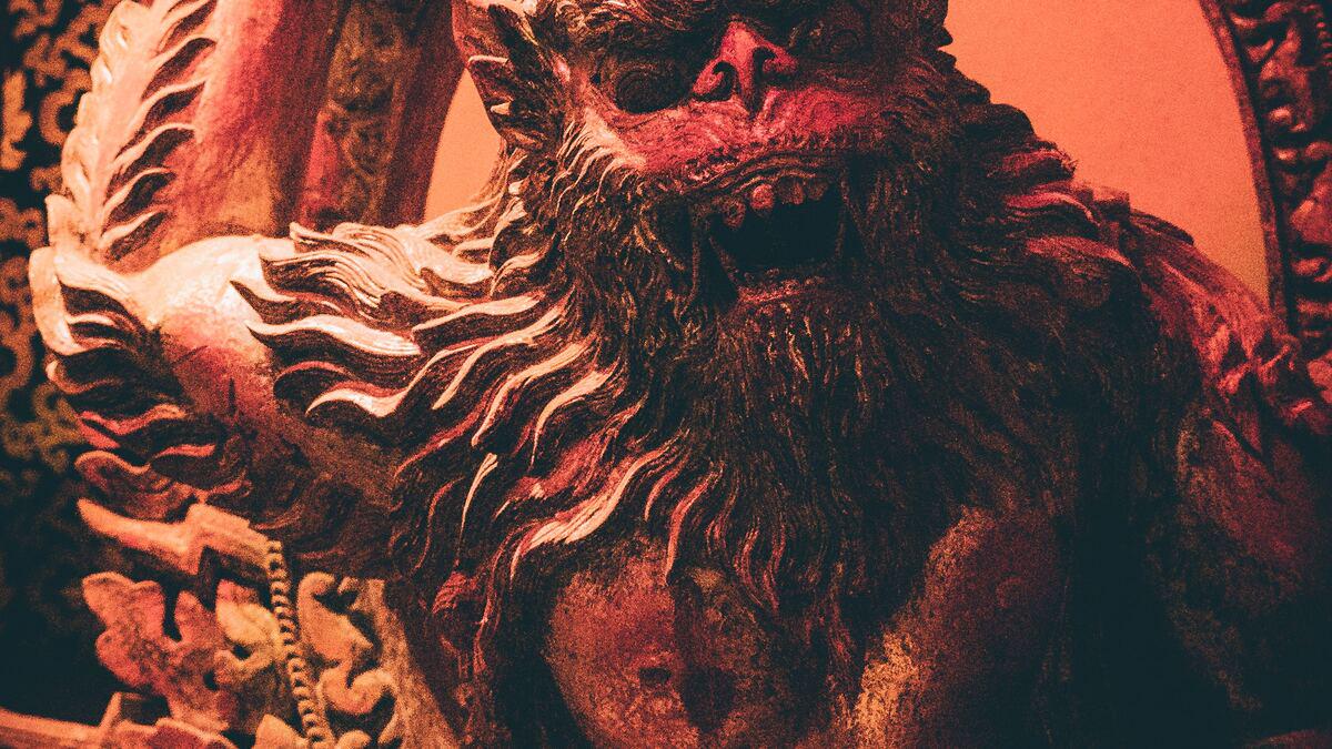 Close-up of a weathered, reddish-brown statue of a mythological creature with a lion-like face and prominent beard. The statue is adorned with ornate carvings and appears to be part of a larger structure.