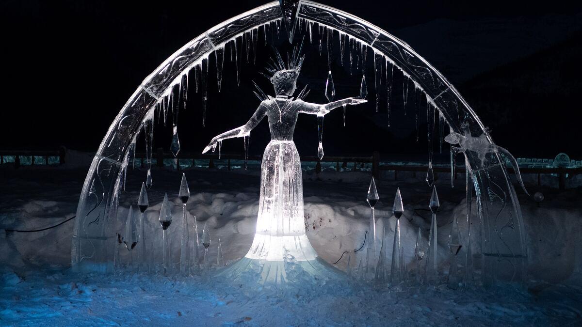 A translucent ice sculpture of a woman with outstretched arms, wearing a crown, and standing beneath an arch adorned with icicles and spear-shaped sculptures, is illuminated against a dark, snowy background. A small ice sculpture of an animal, possibly a lizard, is visible on the right side of the arch.
