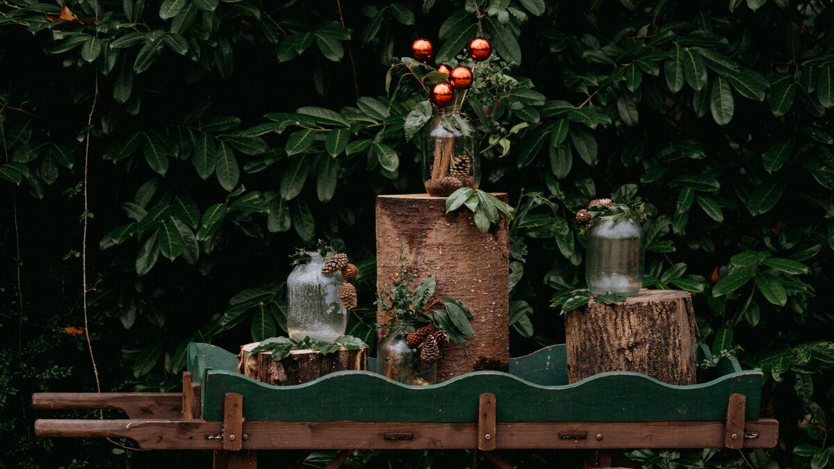 A rustic holiday display sits on a green wooden cart, featuring mason jars filled with pinecones, greenery, and orange ornaments, all arranged on various sized tree stumps against a backdrop of lush foliage. The scene has a natural, woodland-inspired aesthetic.