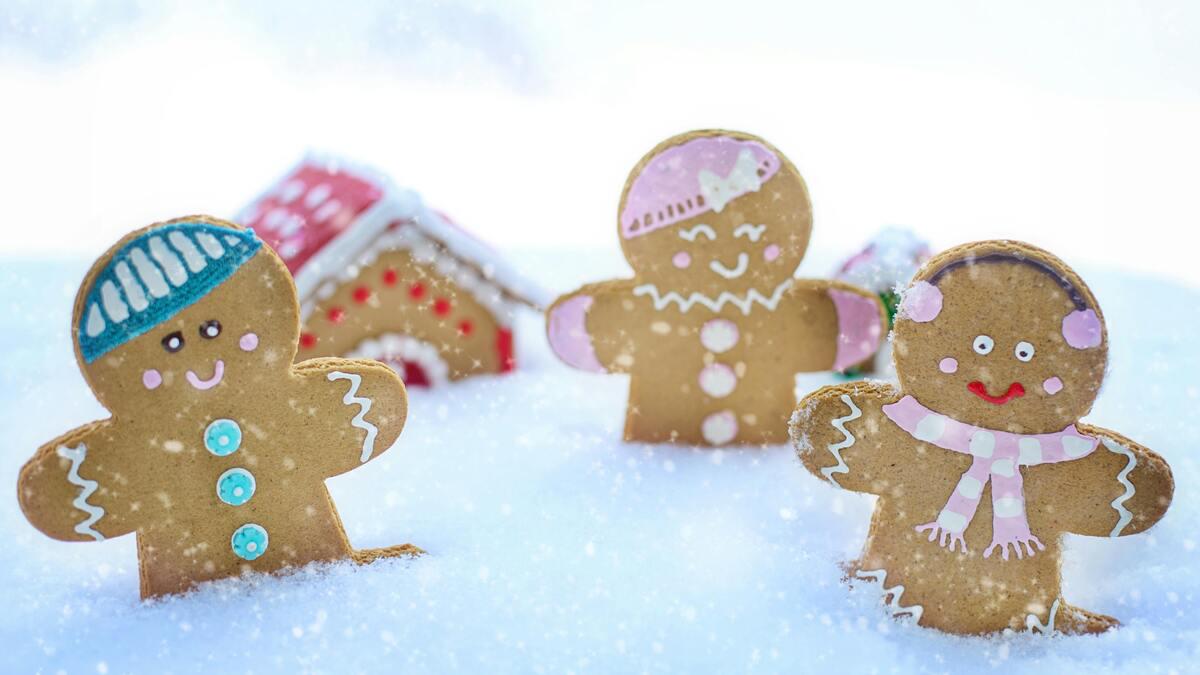Three decorated gingerbread people stand in the snow, with a blurred gingerbread house in the background. The gingerbread figures are adorned with colorful frosting, including hats and scarves.