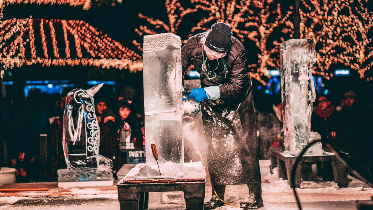 At a winter festival, an ice sculptor in protective gear uses a power tool to carve a block of ice in front of a crowd, with festive lights in the background and a finished ice sculpture of a penguin to the left.