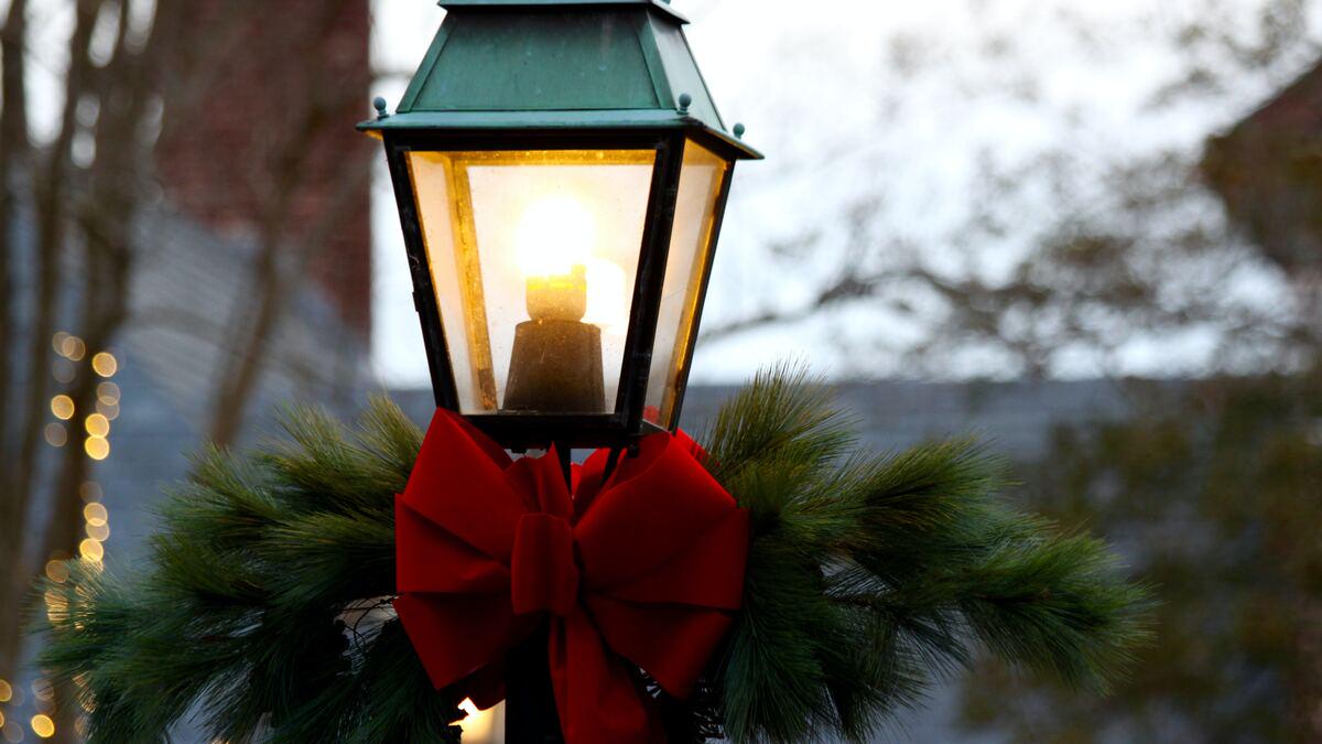 A glowing street lamp is decorated for the holidays with a large red bow and green pine branches. Soft, blurred bokeh lights and buildings can be seen in the background.