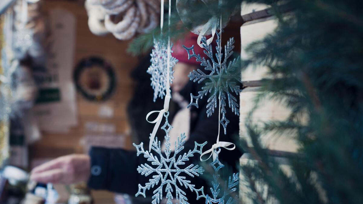 A close-up of hanging silver snowflake ornaments with white ribbons at a Christmas market, with a blurred person in a red hat and wooden stall in the background. The edge of a pine tree branch is visible on the right.