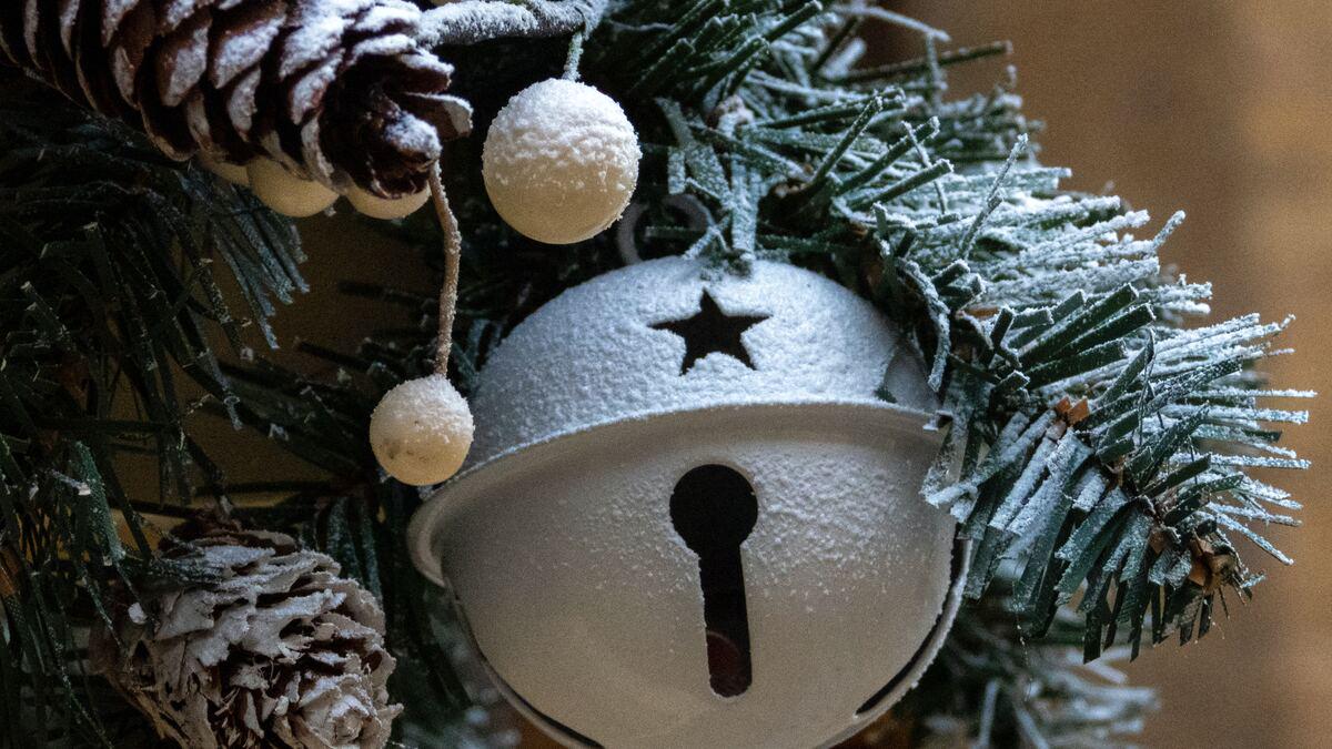 Close-up of snow-dusted Christmas ornaments on a tree branch, including a metal bell with a star cut-out, white balls, and pinecones. The scene evokes a festive, winter holiday mood.