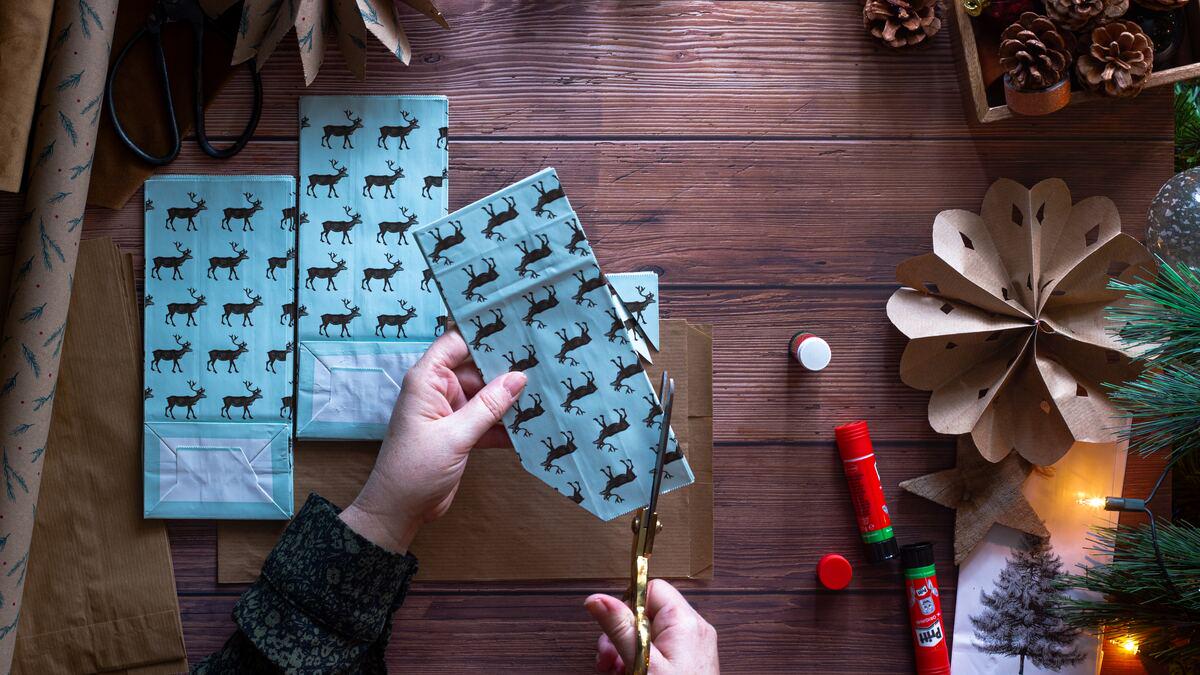 Overhead shot of hands cutting light blue reindeer-patterned paper with gold scissors, surrounded by brown paper bags, glue sticks, pine cones, paper decorations, and Christmas tree sprigs on a wooden surface, suggesting a festive crafting scene.