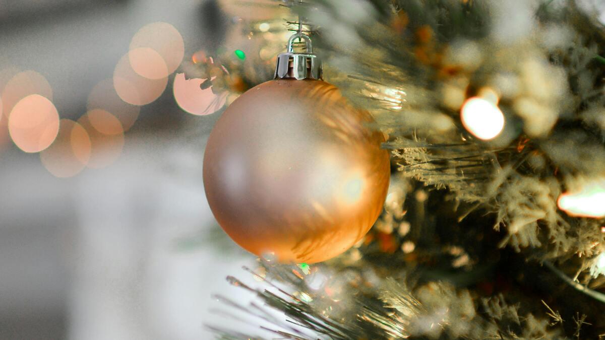A frosted Christmas tree is decorated with a large, gold ornament, while bokeh lights shimmer in the background.