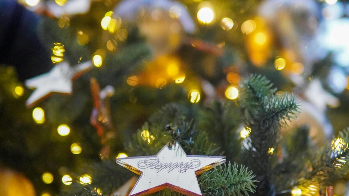 Close-up of a decorated Christmas tree with warm, bokeh-effect lights. A large star ornament with stylized text hangs on a branch in the foreground.