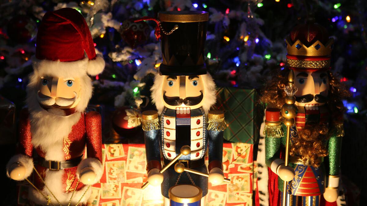 Three decorative nutcrackers, including one dressed as Santa Claus, stand in front of gifts and a Christmas tree with colorful lights. The central nutcracker wears a blue uniform and is holding drumsticks.