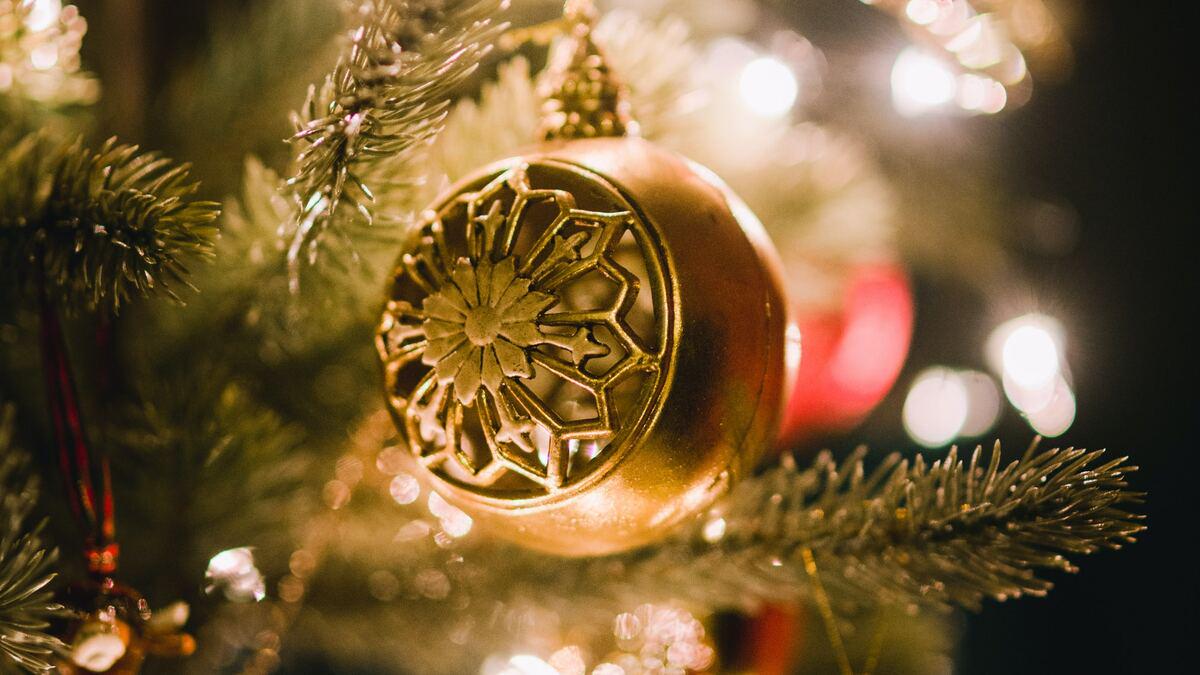 Close-up of a gold Christmas ornament with a snowflake design hanging on a tree with lights in the background. The focus is on the ornament, highlighting its texture and decorative details.