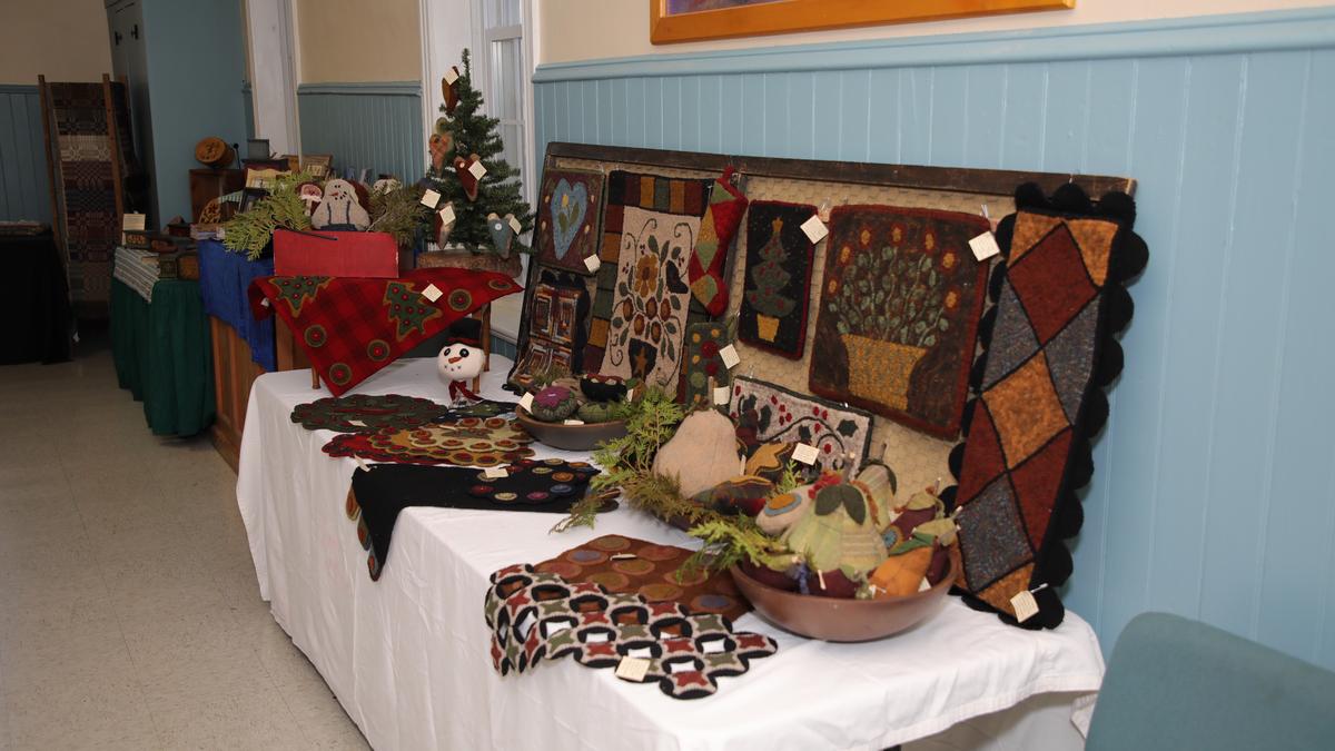 A craft fair display features handmade, hooked rugs and decorative items on tables covered with white cloths. The rugs showcase various patterns and motifs, including Christmas themes, floral arrangements, and geometric designs, creating a festive and artisanal atmosphere.