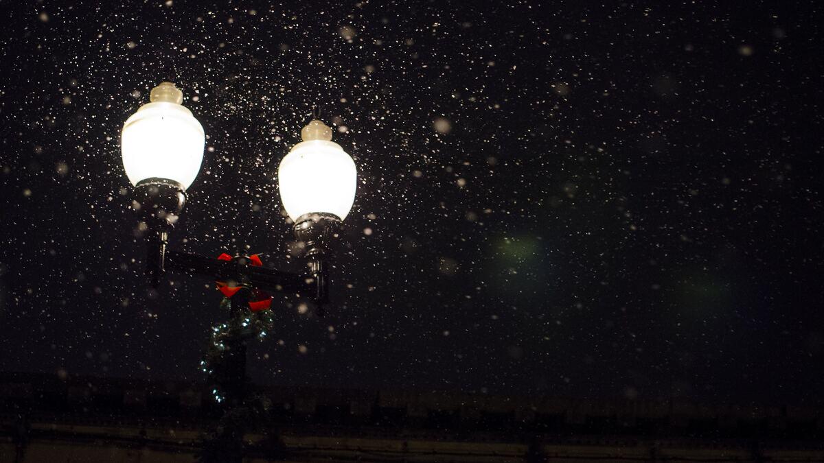 Two illuminated street lamps stand out against a dark night sky filled with falling snow. The lamp post is decorated with greenery, red bows, and small white lights.