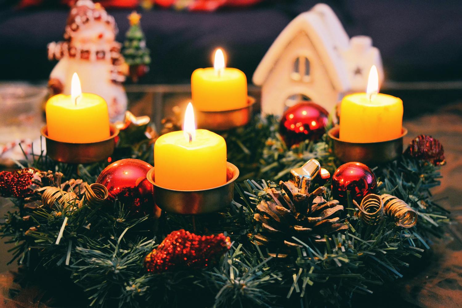 A festive Christmas centerpiece is displayed, featuring four lit yellow candles in metal holders nestled in a wreath of pine needles, with red ornaments, pinecones, and a small ceramic house. A snowman figure is partially visible in the background.