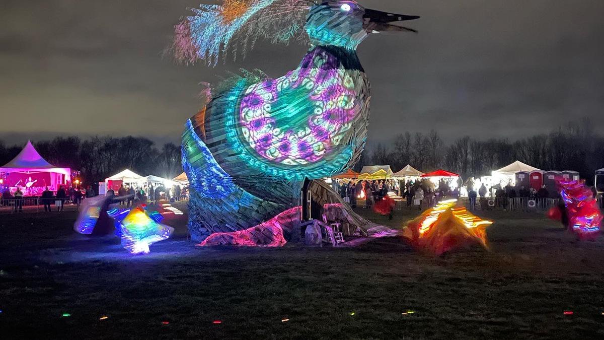 A night scene shows a large, bird-shaped sculpture illuminated with colorful patterns, standing in a field with tents, lights, and people gathered for an event. Smaller illuminated figures are in the foreground, and a band plays on a stage in the distance.