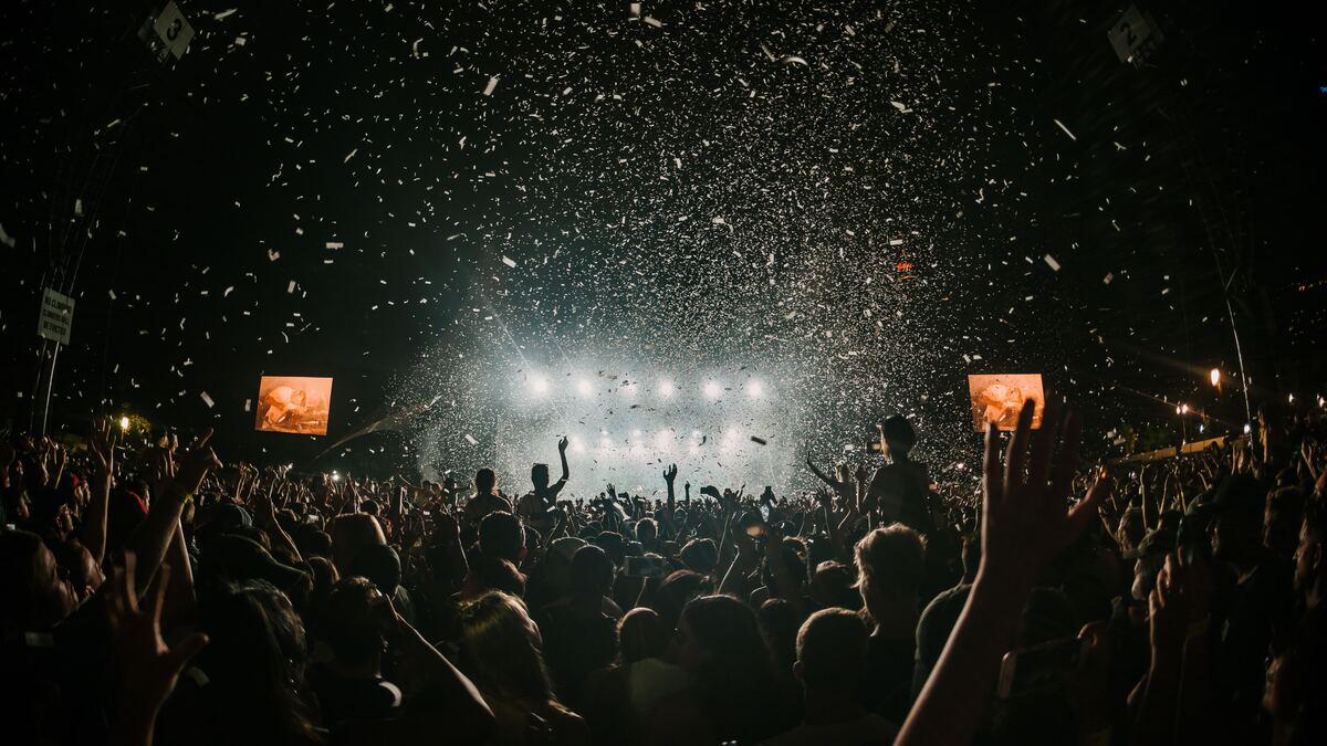 A large crowd of people with raised hands are silhouetted in a dark outdoor space, illuminated by stage lights and falling confetti. Screens are visible on either side of the stage.