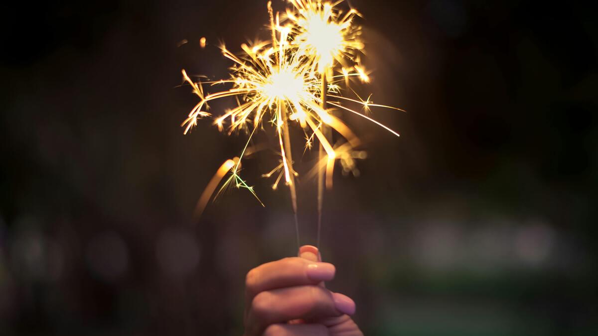 A hand holds two lit sparklers that emit bright sparks, set against a blurred, dark background. The scene evokes a celebratory or festive atmosphere.