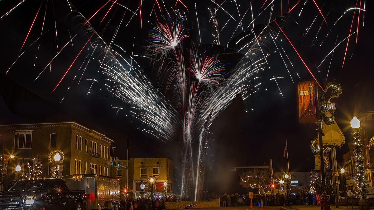 Fireworks explode over a small town square at night, with building facades decorated for the holidays, street lights adorned with wreaths, and a crowd of people watching below. Colorful bursts and streaks of light fill the sky above the lit buildings.