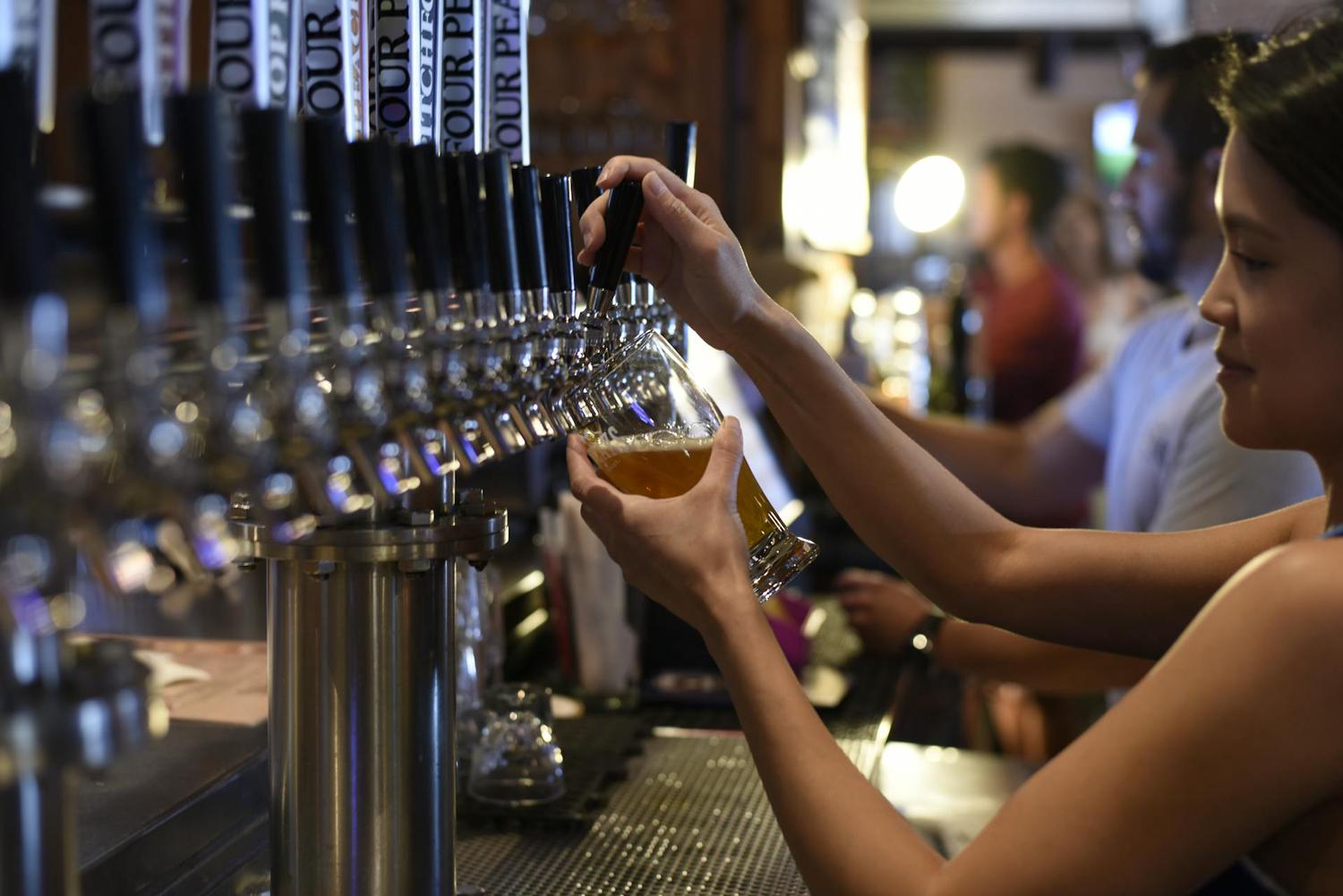 A woman fills a glass with beer from a tap system at a bar. Several other bar patrons are blurred in the background.