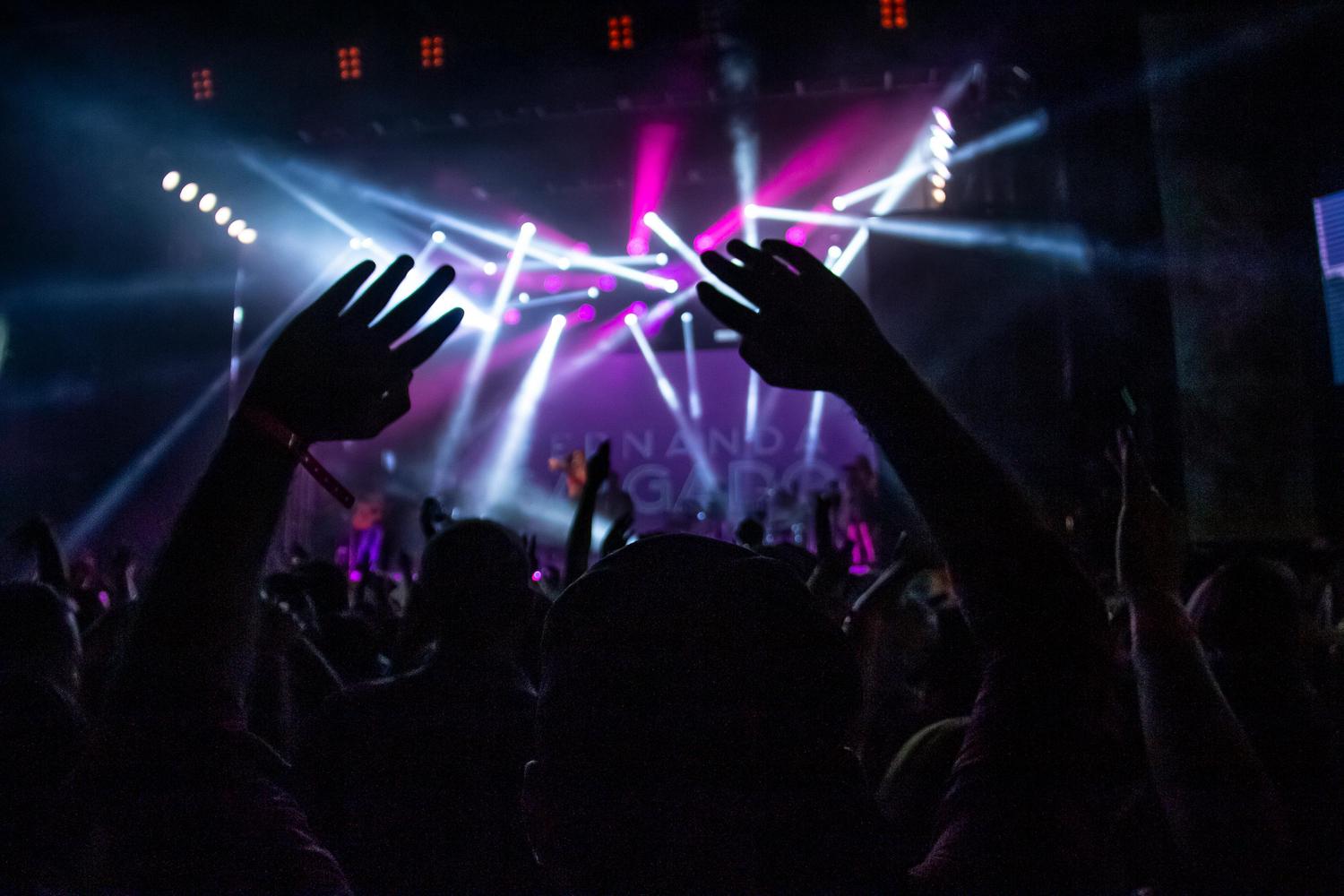 A crowd of people are silhouetted against a stage with bright, white and pink stage lights. Arms are raised in the air, suggesting excitement and engagement with a live performance.