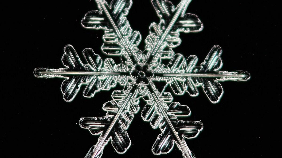 Detailed macro view of a symmetrical snowflake against a black background, showcasing its intricate crystalline structure. The snowflake's delicate, geometric pattern is highlighted with subtle iridescent details.