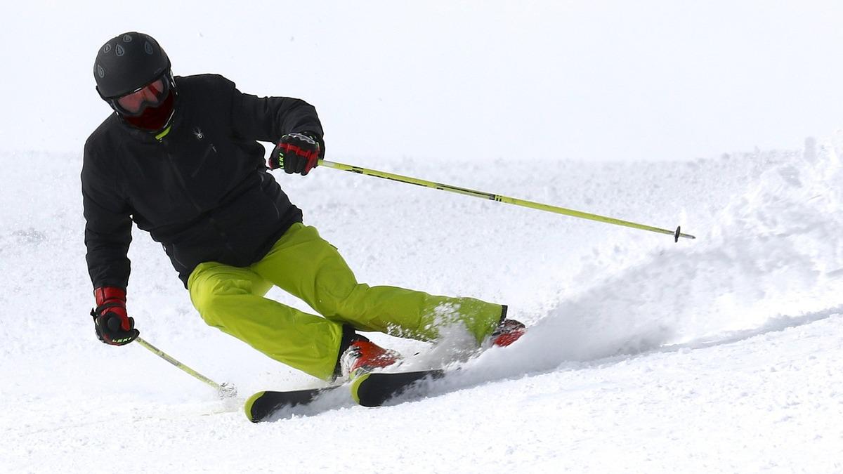 A skier in a black jacket, neon green pants, and a helmet carves a turn in the snow, using ski poles for balance. The bright colors of the clothing contrast with the white snow.