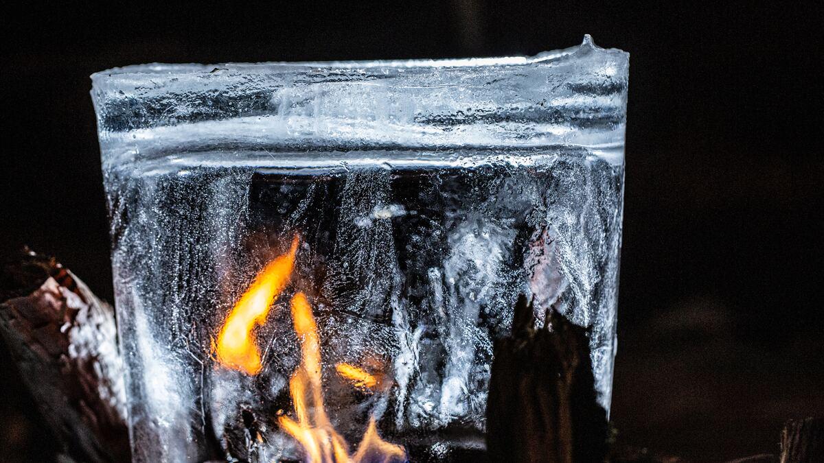 A block of ice stands in front of a fire, reflecting the orange flames within its transparent structure against a dark background. The ice shows textural details and melting areas.