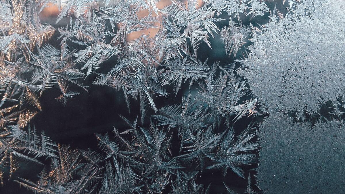 Close-up of frost patterns on a window, resembling delicate ferns with intricate crystalline structures. Part of the glass is completely covered in a dense, frosted layer.