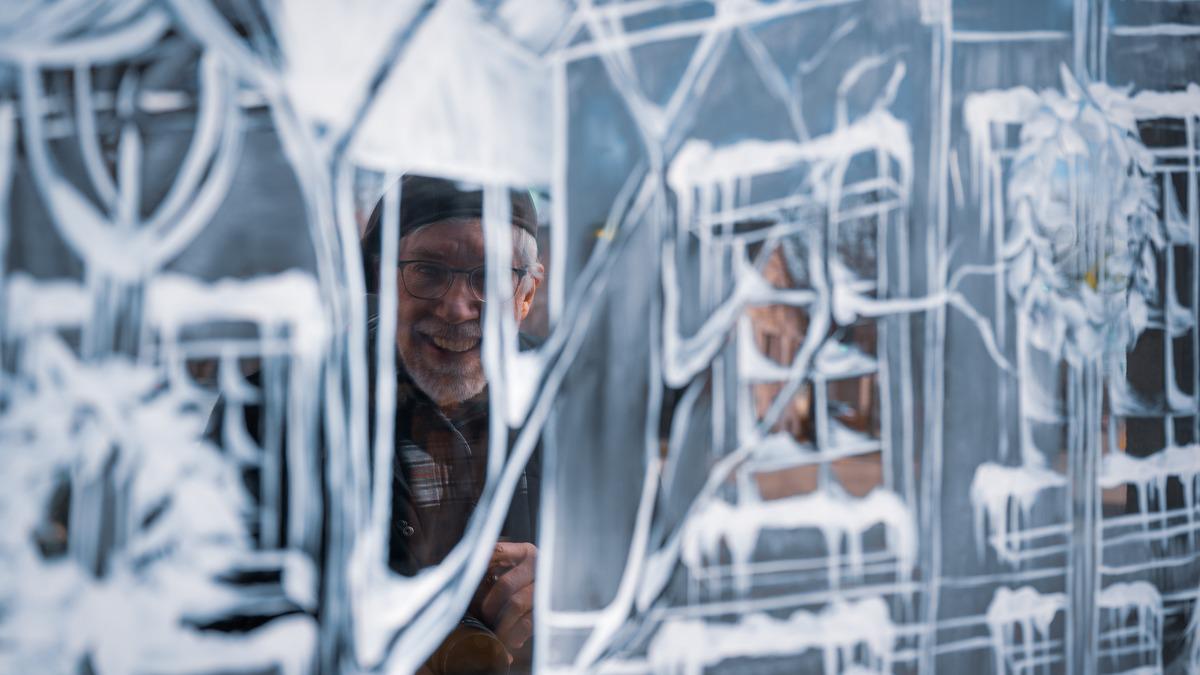 A man with glasses and a hat smiles from behind a window display decorated with winter scenes in white paint. The snowy designs include buildings, trees, and icicles.
