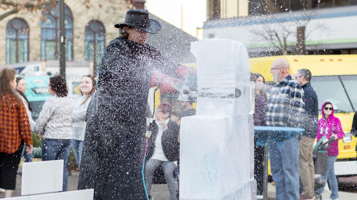 A person wearing a black hat and coat is using a power tool to sculpt a large block of ice in a public space, with spectators watching nearby. Ice shavings are flying from the sculpture as it is being worked on.