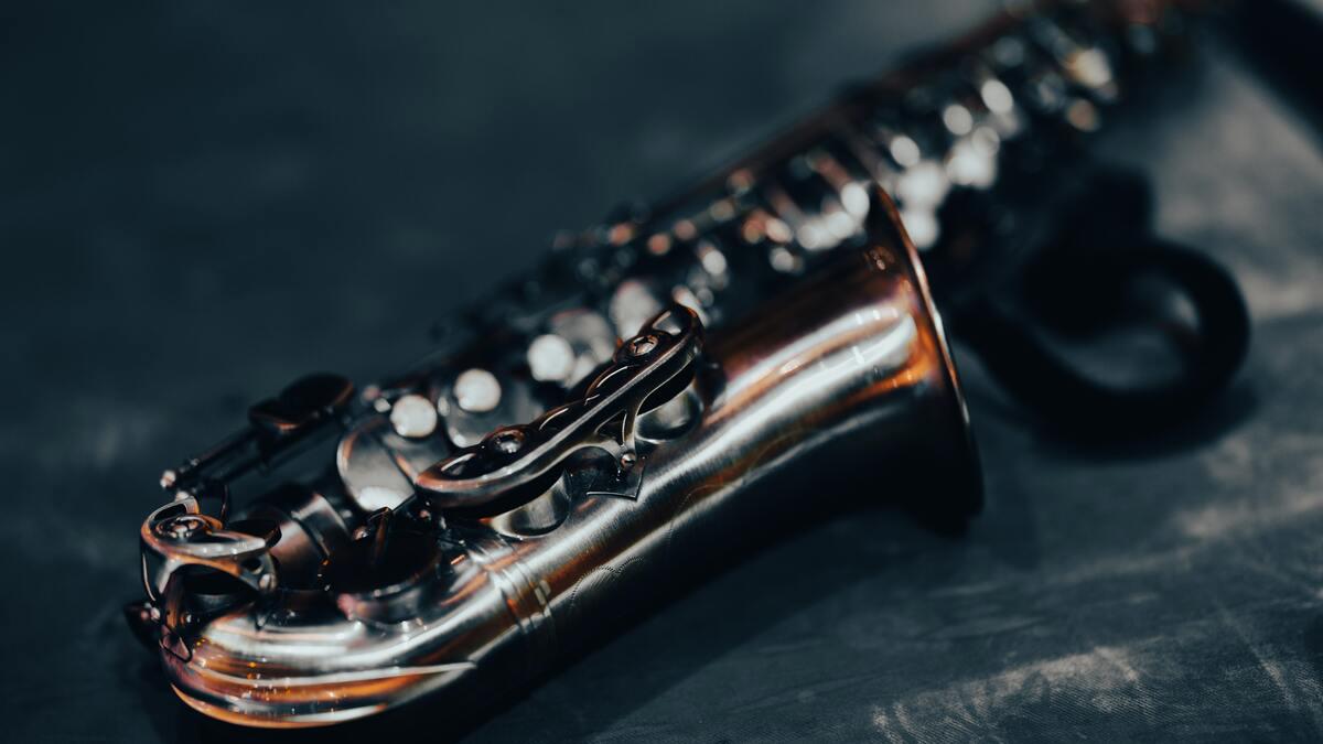 Close-up of a silver saxophone lying on a textured dark surface, with the bell and keys in focus, and the body extending into the blurred background. The lighting highlights the instrument's curves and the complexity of its mechanism.